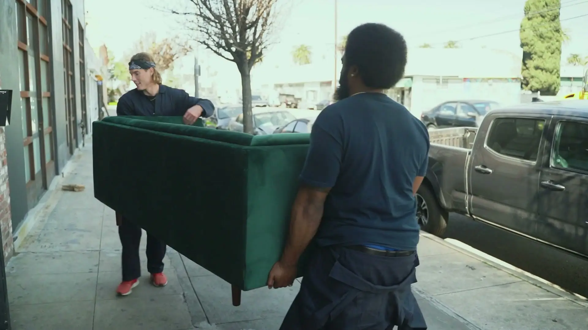 Two workers in navy shirts carrying a green couch along a sidewalk toward a pickup truck